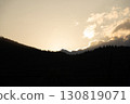 Golden sunlight peeks over mountain peaks at dusk, illuminating clouds and silhouetting trees in the valley 130819071
