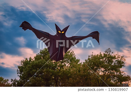 Dark bat-shaped kite with glowing yellow eyes flying against dramatic evening sky over trees, creating a spooky Halloween atmosphere Dark bat-shaped kite with glowing yellow eyes flying against dramatic evening sky over trees, creating a spooky Halloween atmosphere 130819907