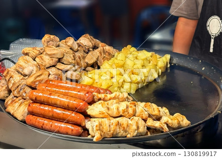Vibrant street food scene with grilled sausages, chunks of chicken, and diced potatoes cooking on large pan, capturing essence of urban take away culinary culture Vibrant street food scene with grilled sausages, chunks of chicken, and diced potatoes cooking on large pan, capturing essence of urban take away culinary culture 130819917