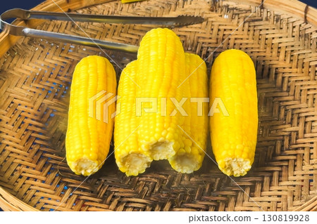 Boiled ears of corn rest on a woven bamboo tray, their bright yellow kernels offering a healthy, quick snack, embodying the simplicity and appeal of traditional street food 130819928