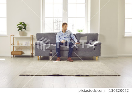 Busy man sitting on sofa at home, working on laptop, and drinking hot coffee from thermos 130820022