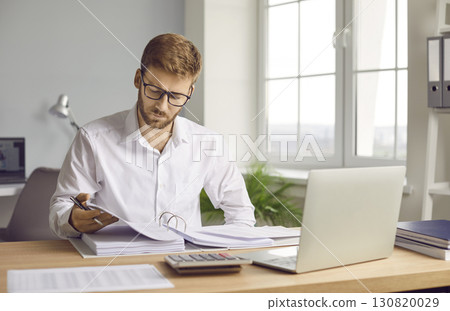 Prtrait of a young businessman looking through paper documents and working at office. 130820029