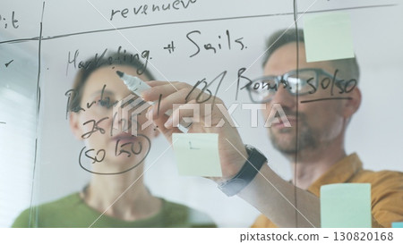 Business professionals reviewing marketing data, sales metrics, and partnership insights on transparent board, collaborating with sticky notes in modern workplace setting Business professionals reviewing marketing data, sales metrics, and partnership insights on transparent board, collaborating with sticky notes in modern workplace setting 130820168