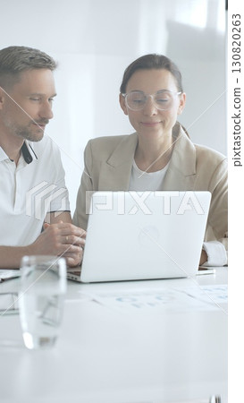 Businessman and adult businesswoman reviewing marketing analytics on laptops, working together strategically in contemporary office workplace. Business people concept 130820263