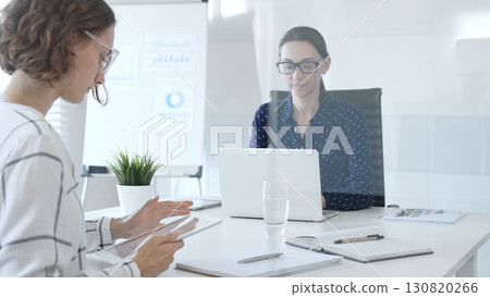 Two businesswomen are sitting at a white table, one of them holding a tablet and talking to her colleague sitting at the laptop, during a business meeting. Business people concept 130820266