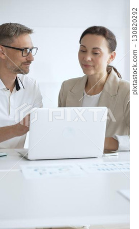Businessman and adult businesswoman reviewing marketing analytics on laptops, working together strategically in contemporary workplace with minimalist interior design and collaborative atmosphere 130820292
