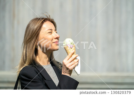Satisfied squinting businesswoman eating ice cream while walking during lunch break in good weather 130820379