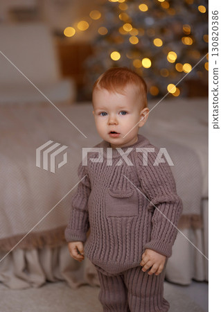 Red-haired baby standing in knitted outfit near bed and Christmas tree, front view 130820386