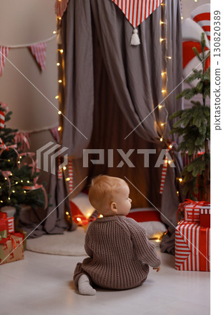 Baby in knitted sweater sitting on floor near Christmas tent and presents, rear view Baby in knitted sweater sitting on floor near Christmas tent and presents, rear view 130820389