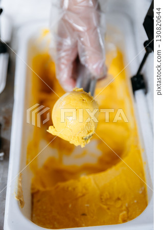 Hand scooping vibrant yellow mango gelato in a container, close-up food preparation Hand scooping vibrant yellow mango gelato in a container, close-up food preparation 130820644