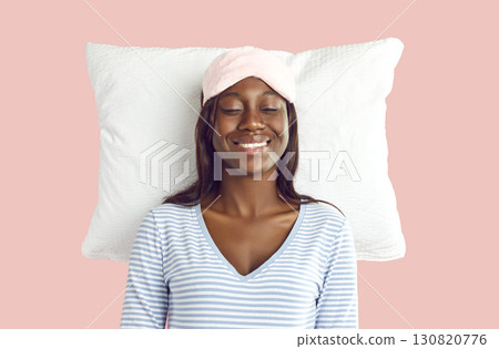 Happy young woman in sleep eye mask isolated on studio pink background lying on white soft pillow. Happy young woman in sleep eye mask isolated on studio pink background lying on white soft pillow. 130820776