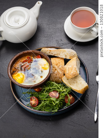 Baked eggs with vegetables in clay bowl, served with ciabatta bread and fresh salad 130821018