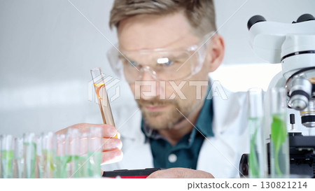 Man researcher with safety glasses is examining lab tube with a orange fluid in a modern laboratory, close-up. Science, microbiology and medicine 130821214