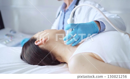 Doctor man wearing blue medical gloves is using ultrasound equipment on neck of female patient lying down for a medical examination Doctor man wearing blue medical gloves is using ultrasound equipment on neck of female patient lying down for a medical examination 130821256