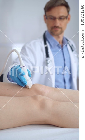 Doctor man with blue protective gloves using ultrasound equipment on the knee of a female patient, analyzing the results on a monitor in the background. Medicine 130821260