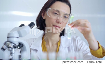 Scientist researcher with protective glasses examining lab tube with a green fluid. Microbiology and science concept Scientist researcher with protective glasses examining lab tube with a green fluid. Microbiology and science concept 130821281