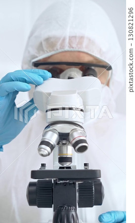 Scientist in ppe analyzing samples with microscope. Close-up of a focused researcher in a lab coat and protective gear in a laboratory setting Scientist in ppe analyzing samples with microscope. Close-up of a focused researcher in a lab coat and protective gear in a laboratory setting 130821296