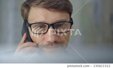 Businessman with glasses speaking on the phone, partially obscured by blurred laptop. Front view and close up portrait 130821322