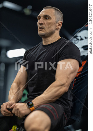 Muscular man sits on a bench in a contemporary gym catching his breath after an intense workout session. The atmosphere is energetic with various gym equipment visible in the background. Muscular man sits on a bench in a contemporary gym catching his breath after an intense workout session. The atmosphere is energetic with various gym equipment visible in the background. 130821647