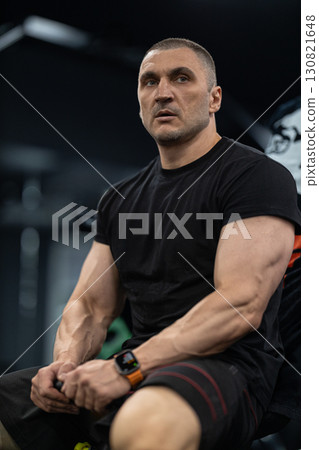 athletic man sits on a bench in a gym taking a moment to rest. He appears focused and determined after completing a challenging workout. The environment suggests a dedicated training space. 130821648