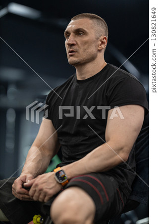 athletic man sits on a bench in a modern gym appearing focused and tired after an intense workout. He is dressed in a black shirt and shorts accompanied by a smartwatch. 130821649