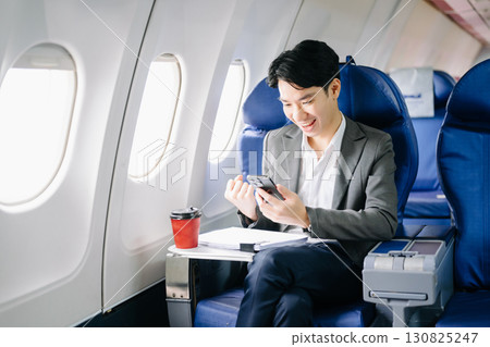 Asian businessman or entrepreneur in a formal suit on an airplane sitting in business class using a phone, computer laptop 130825247