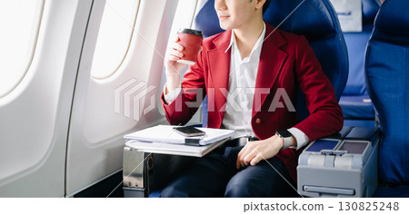 Asian businessman or entrepreneur in a formal suit on an airplane sitting in business class using a phone, computer laptop 130825248