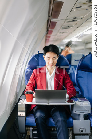 Asian businessman or entrepreneur in a formal suit on an airplane sitting in business class using a phone, computer laptop 130825252