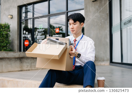 Businessman sitting outside the office with a box of belongings, reflecting on job loss. Capturing the emotional impact and reality 130825473