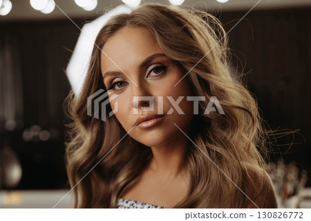 Portrait of attractive young woman with wavy hair. Behind woman there is mirror and lights of beauty salon. Model looks expressively at camera. Close-up Portrait of attractive young woman with wavy hair. Behind woman there is mirror and lights of beauty salon. Model looks expressively at camera. Close-up 130826772