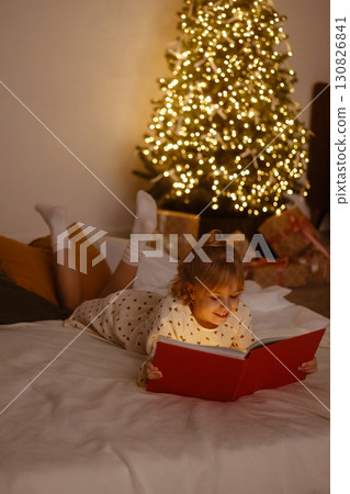 Little girl lying on bed reading red book near Christmas tree with lights and gifts 130826841