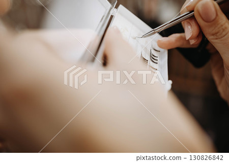 Eyelash extensions. Hand of makeup artist takes eyelashes with tweezers for subsequent gluing of extensions Eyelash extensions. Hand of makeup artist takes eyelashes with tweezers for subsequent gluing of extensions 130826842