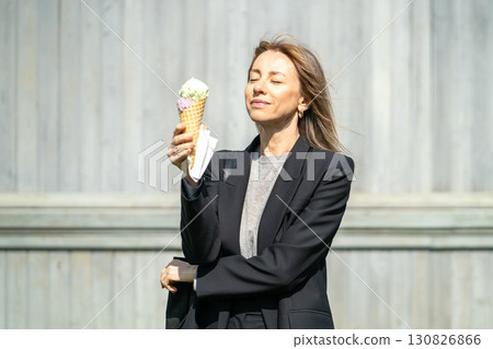 Happy satisfied businesswoman savoring and enjoying ice cream cone after work on urban streets Happy satisfied businesswoman savoring and enjoying ice cream cone after work on urban streets 130826866