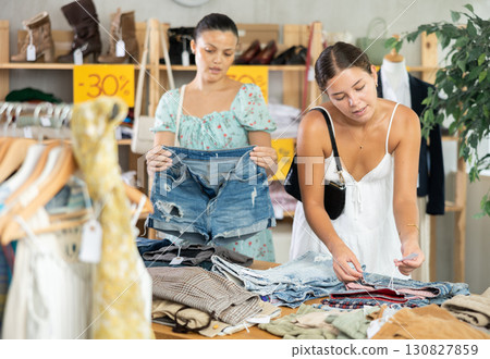 Two young women carefully choosing fashionable jeans or shorts in clothing store 130827859