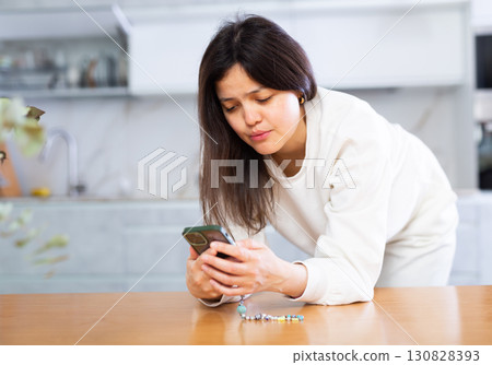 Upset young brunette standing in home kitchen with smartphone 130828393