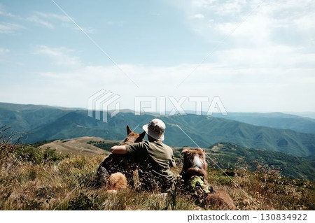Woman with two dogs German and Australian Shepherd sitting on a mountain meadow in Stara Planina, Serbia, enjoying the scenic panoramic view of green ridges under a bright sky. Hiking with pets Woman with two dogs German and Australian Shepherd sitting on a mountain meadow in Stara Planina, Serbia, enjoying the scenic panoramic view of green ridges under a bright sky. Hiking with pets 130834922