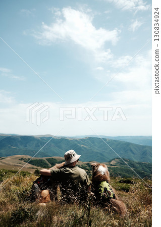 Woman with two dogs German and Australian Shepherd sitting on a mountain meadow in Stara Planina, Serbia, enjoying the scenic panoramic view of green ridges under a bright sky. Hiking with pets 130834924