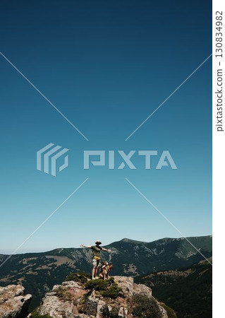 Female hiker with arms wide open standing on a rocky mountain top of Stara Planina with german shepherd dog, nature and freedom concept. Travel and hiking with pet concept 130834982