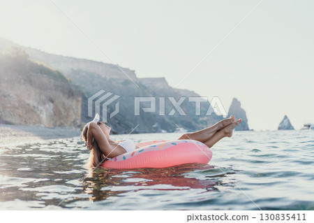 Woman, relaxing, ocean. Serene woman floating on a pink donut in the sea near rocky cliffs, copy space. 130835411