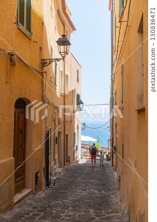 People walk down a narrow Mediterranean alley, surrounded by charming buildings, towards a scenic ocean view. The serene scene captures the essence of coastal village life. Sardinia  130836471