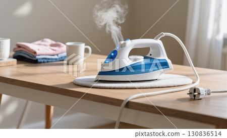 Steam Iron on a Wooden Table With Freshly Folded Laundry in a Sunlit Room 130836514