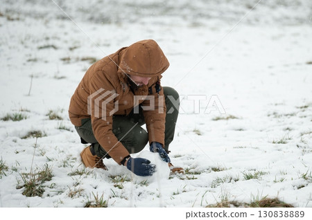 A person crouches in the snow, carefully shaping a small snowball. A person crouches in the snow, carefully shaping a small snowball. 130838889