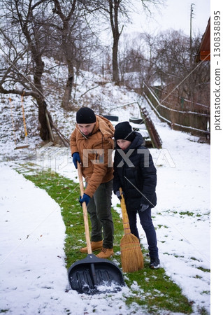 Father and son teamwork: clearing snow from their yard with a shovel and broom. A winter chore done together. 130838895