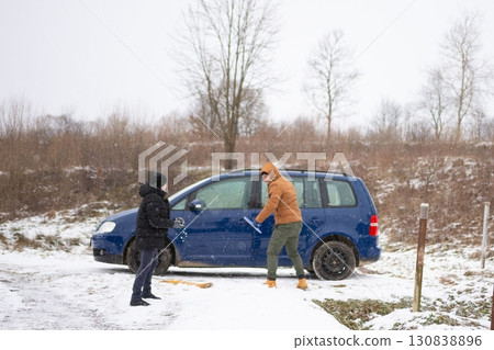 Two people clearing snow from a car in a snowy landscape. One is using an ice scraper while the other waits. Two people clearing snow from a car in a snowy landscape. One is using an ice scraper while the other waits. 130838896