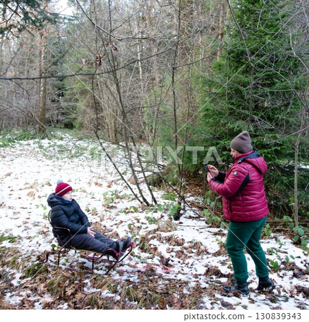 A child sits on a sled in a snowy forest while an adult takes a photo. A child sits on a sled in a snowy forest while an adult takes a photo. 130839343