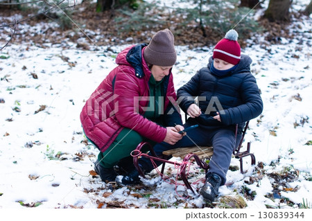 A woman and child use a phone together in a snowy forest, near a small wooden sled. A woman and child use a phone together in a snowy forest, near a small wooden sled. 130839344