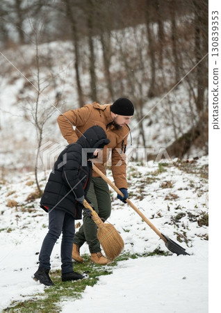 Father and son teamwork: cleaning snow together in a wintery landscape using a broom and shovel. Father and son teamwork: cleaning snow together in a wintery landscape using a broom and shovel. 130839353