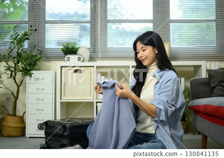 Young Asian woman folding clothes and packing suitcase while preparing for travel in cozy living room Young Asian woman folding clothes and packing suitcase while preparing for travel in cozy living room 130841135
