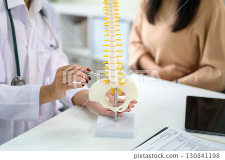 Doctor using a spine model to explain lower back issues to patient during medical consultation Doctor using a spine model to explain lower back issues to patient during medical consultation 130841198