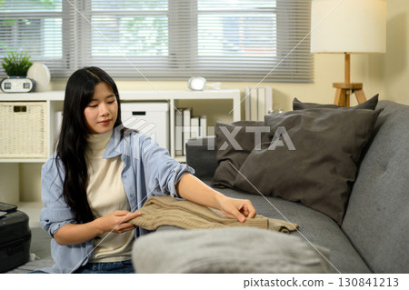 Young Asian woman folding clothes and packing suitcase while preparing for travel in cozy living room Young Asian woman folding clothes and packing suitcase while preparing for travel in cozy living room 130841213